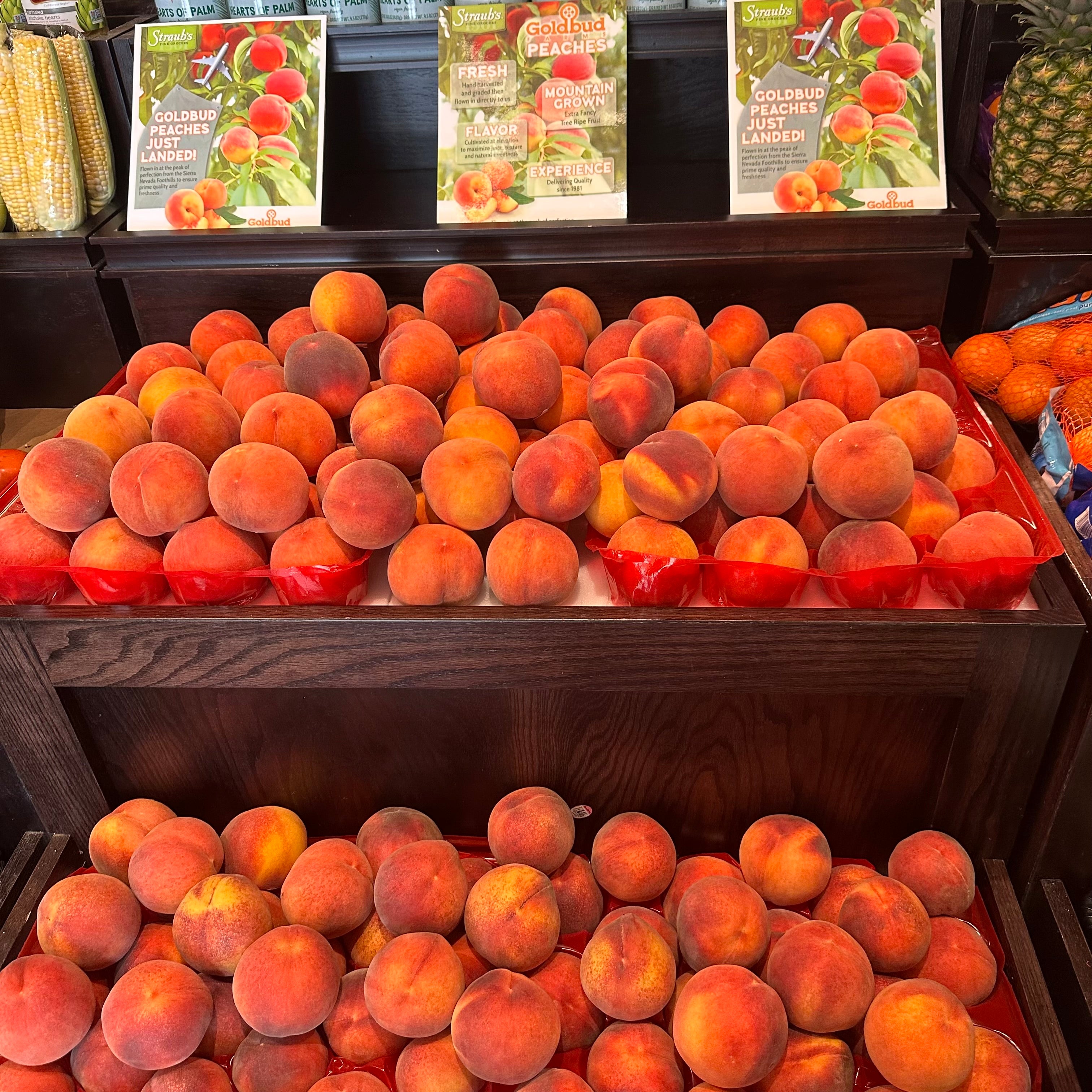 Fresh peaches displayed on dark wooden market shelves, with printed fruit signboards in the background.