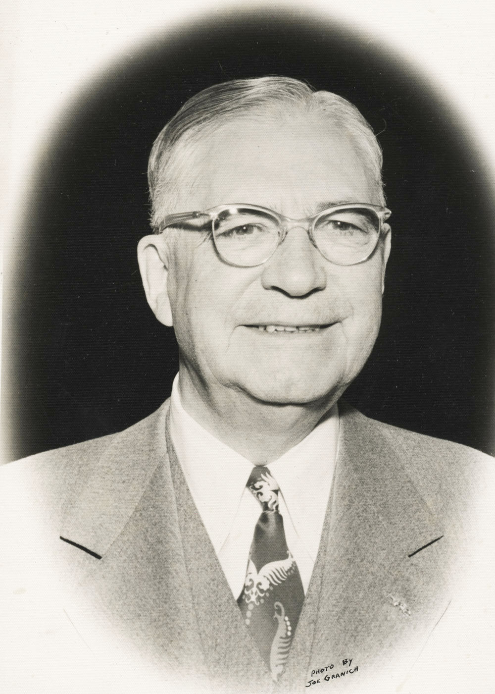 Black and white portrait of a man wearing glasses and a suit with a tie.