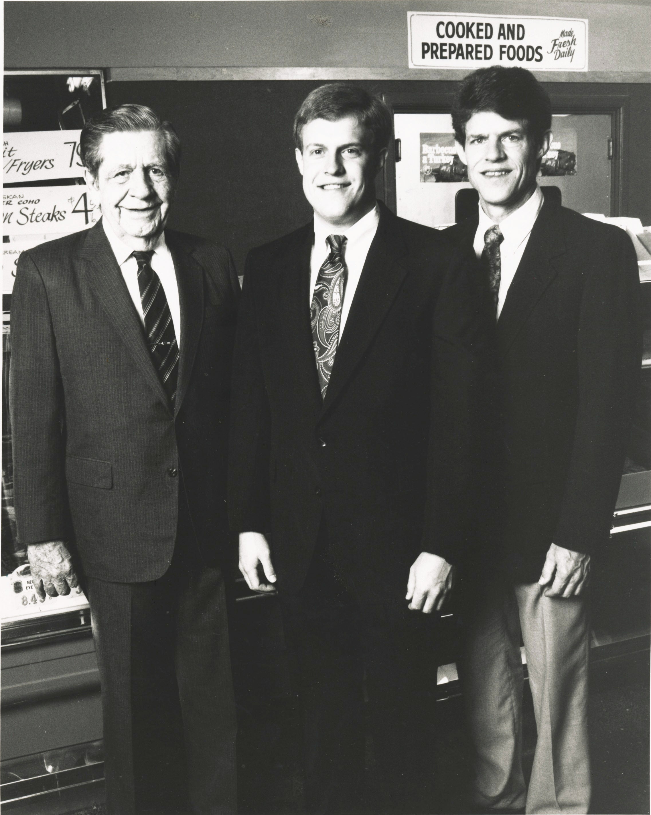 Three men in suits standing in front of a store display with 'Cooked and Prepared Foods' sign.
