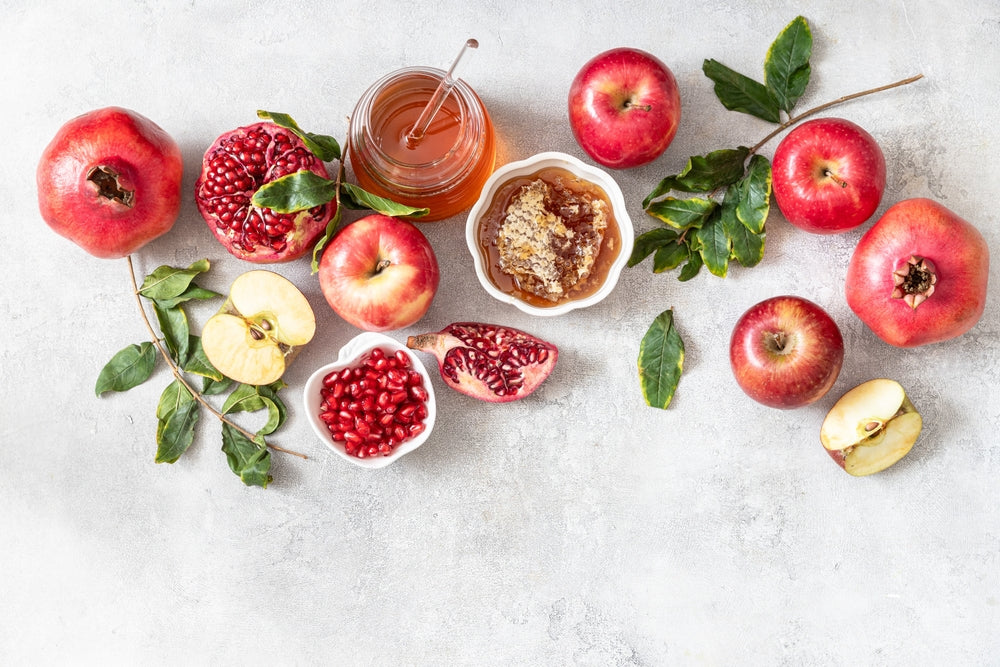 A styled flat-lay of honey and pomegranate ingredients on a light gray textured surface, including whole and sliced red apples, whole and halved pomegranates, green leaves, and two small white bowls containing honey and pomegranate seeds.